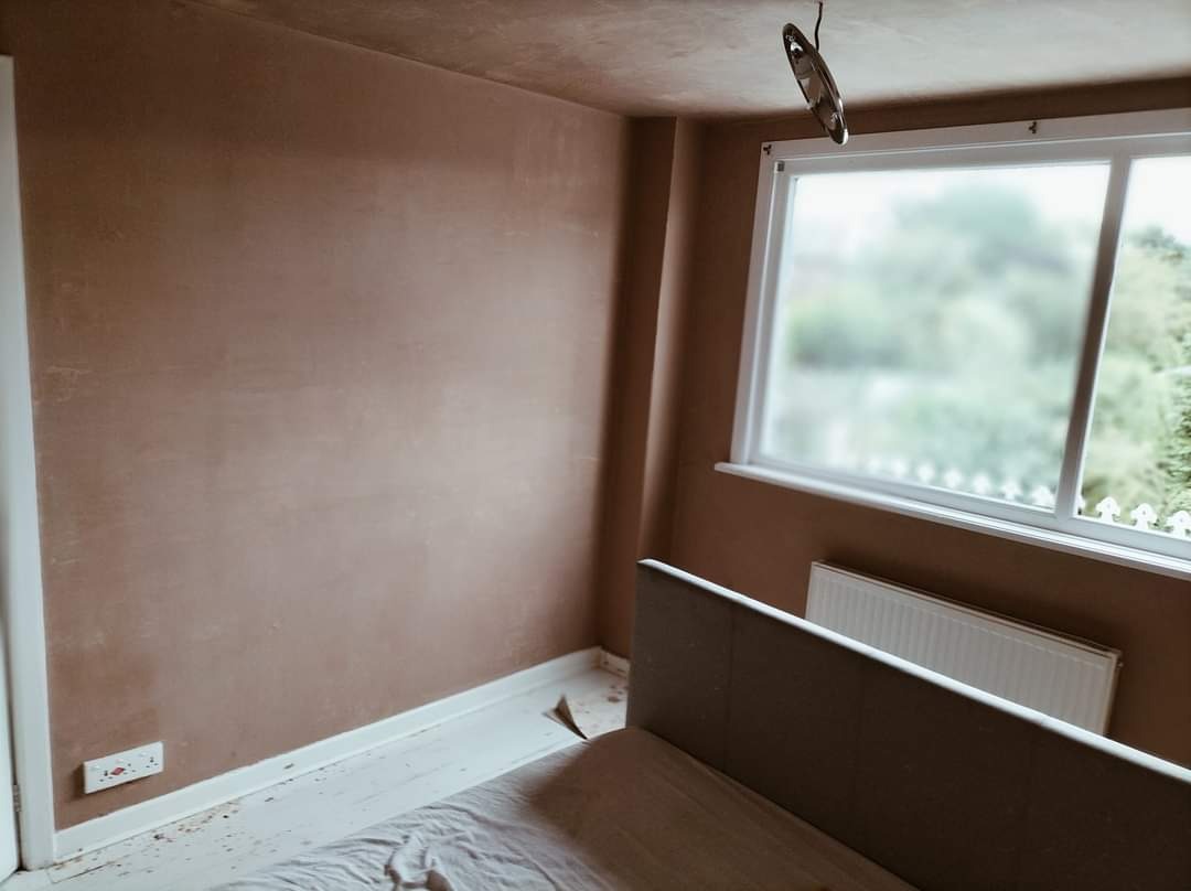 Pokój po nałożeniu gładzi szpachlowej na ściany, z widocznym fragmentem łóżka przykrytego folią ochronną, białe listwy przypodłogowe i okno z dekoracyjną naklejką. Na suficie wisi garnek.