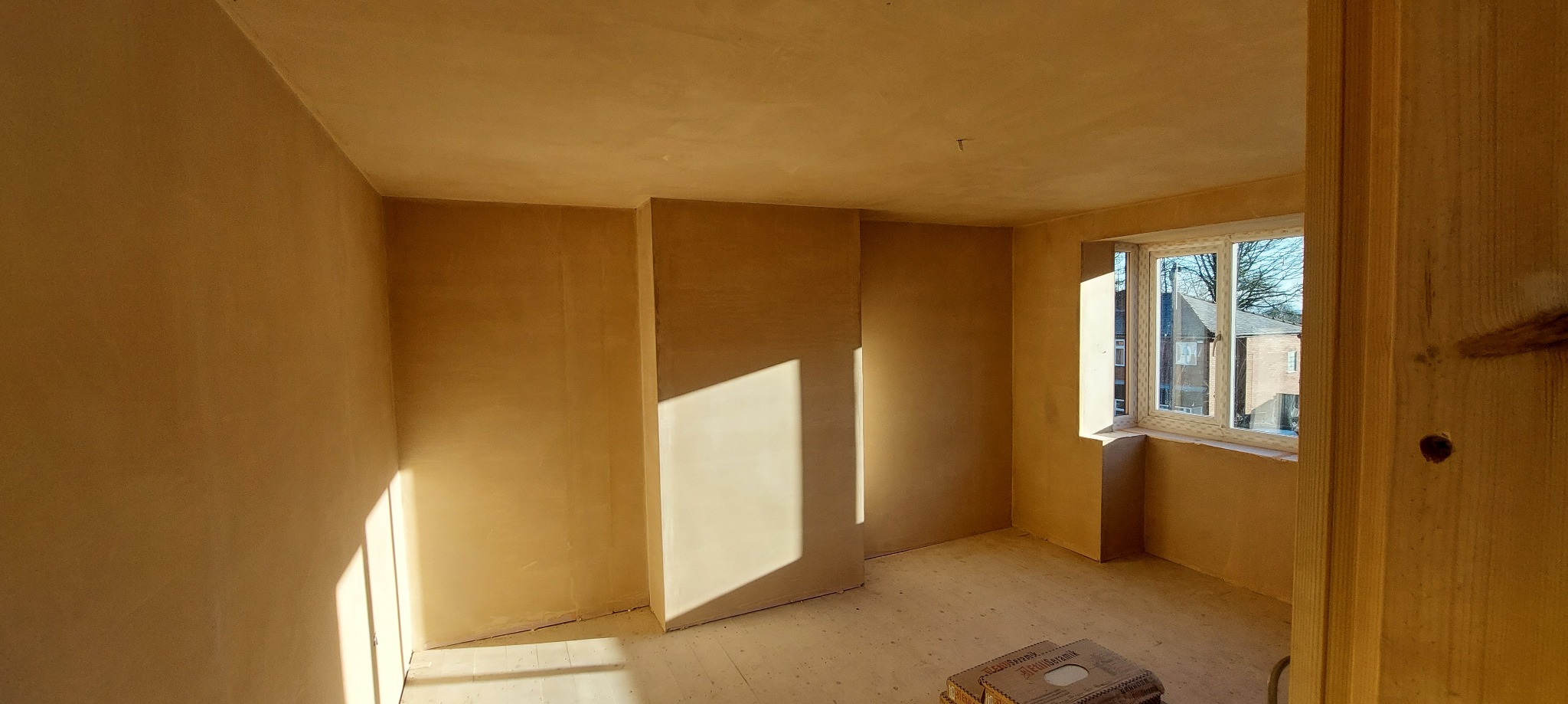 Pokój po nałożeniu gładzi szpachlowej, widoczne ślady szlifowania, jasne światło słoneczne padające przez okno, kartony z materiałem na podłodze.