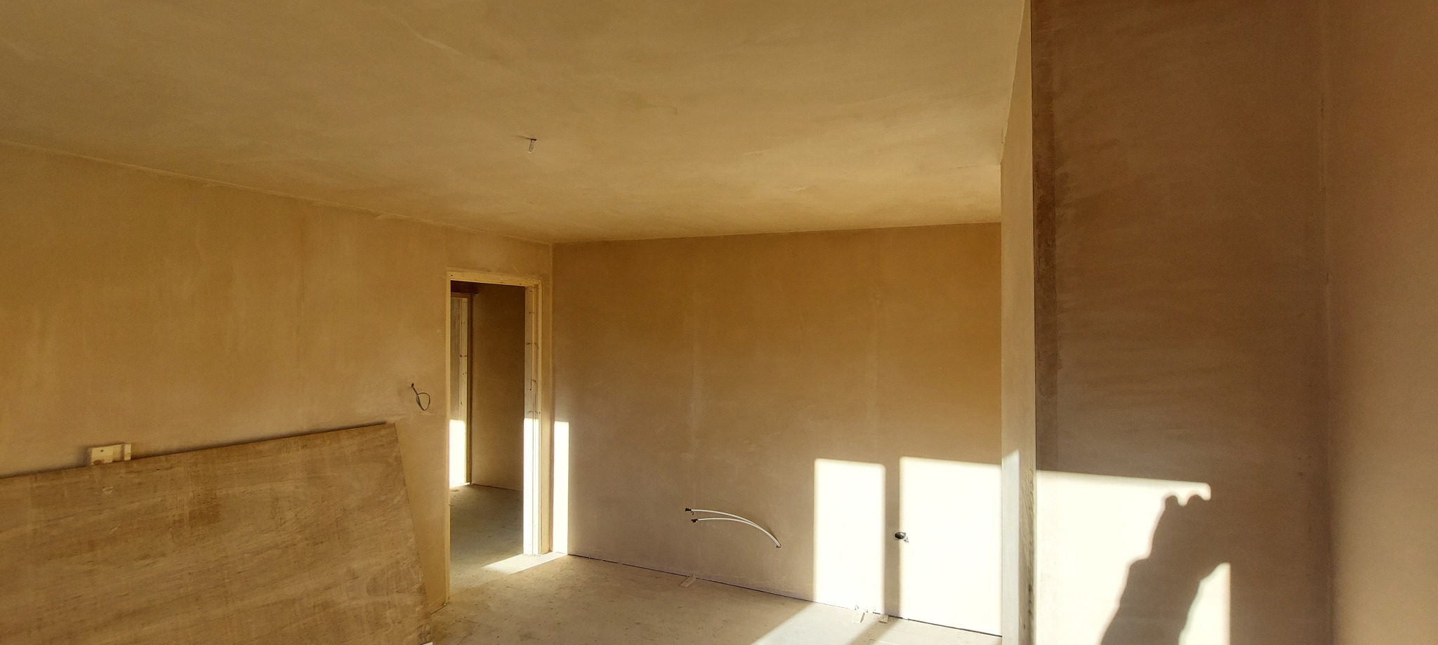 Pokój w trakcie prac wykończeniowych: ściany pokryte świeżo nałożoną, jasną szpachlą, widoczne przewody instalacji elektrycznej, drewniana płyta oparta o ścianę, światło słoneczne wpadające przez...