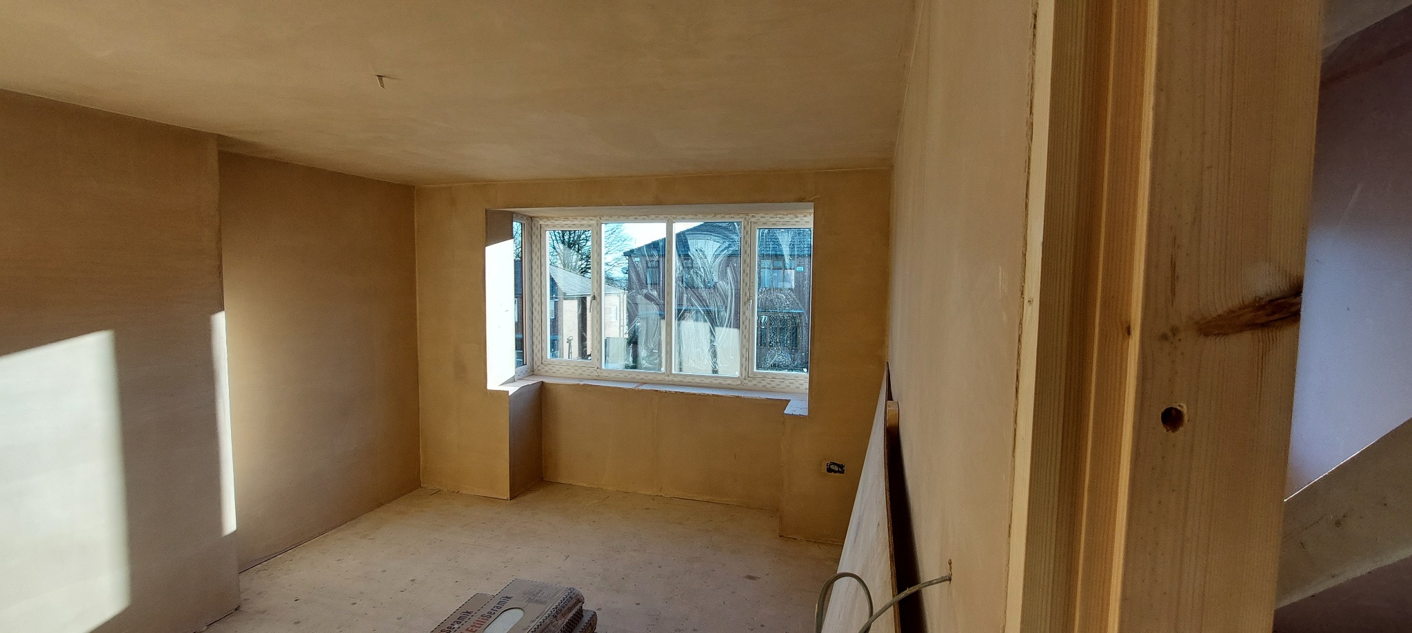 Pokój po nałożeniu gładzi szpachlowej na ścianach i suficie, widoczne okno z widokiem na zabudowę mieszkalną, w rogu pomieszczenia płyta kartonowo-gipsowa oparta o ścianę.