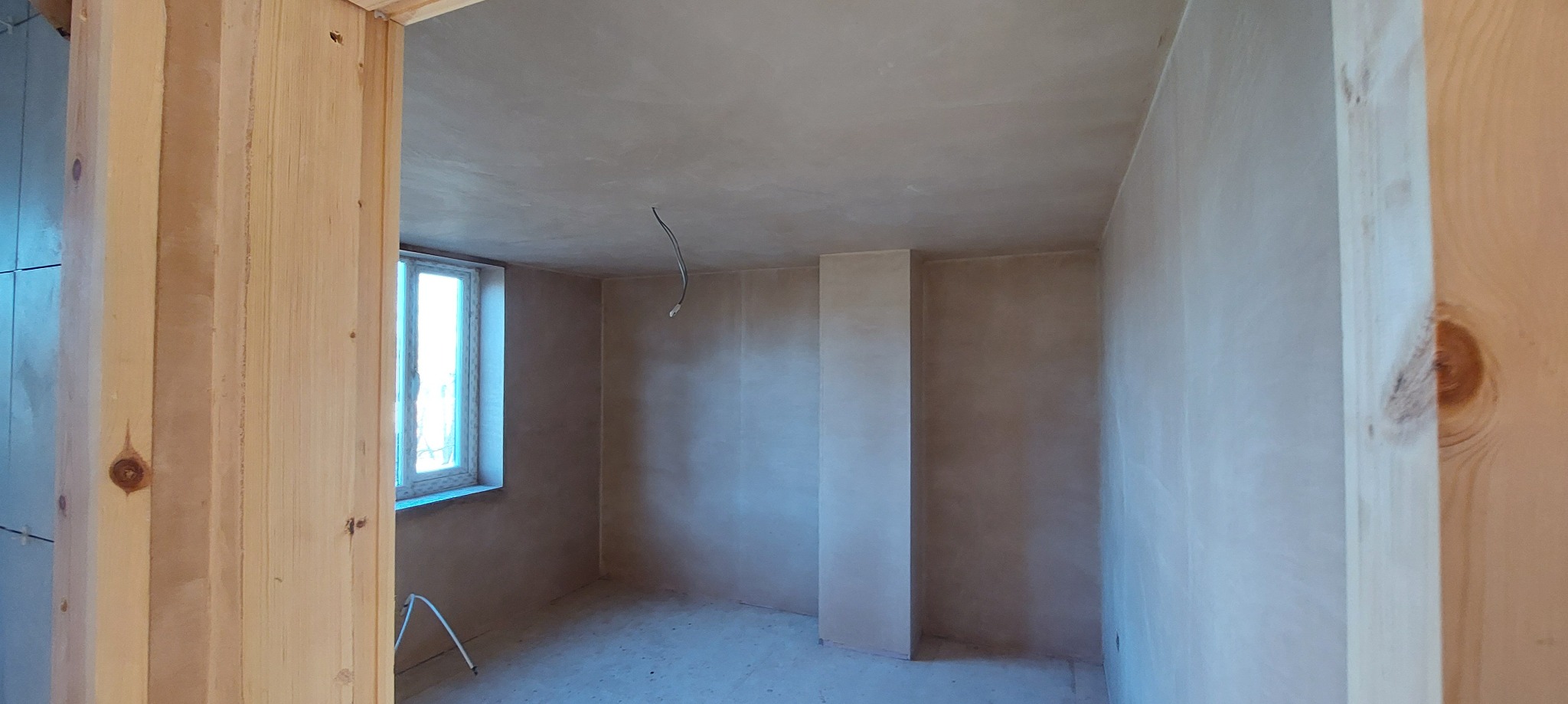 Pokój w trakcie przygotowania do malowania, ściany pokryte szpachlą, widoczne drewniane futryny drzwi i okno z parapetem, wystający kabel z sufitu i metalowy stojak pod oknem.