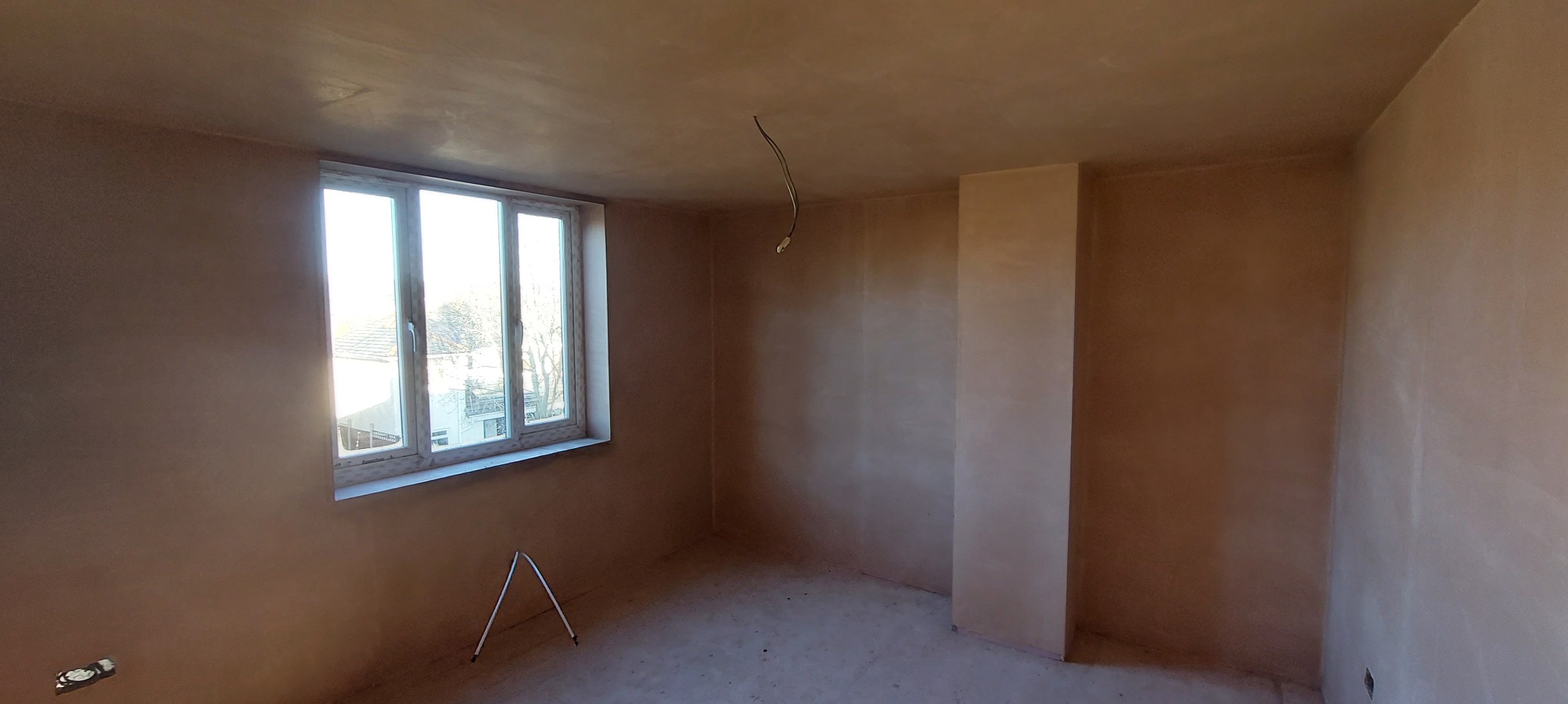 Pokój po nałożeniu gładzi szpachlowej, widoczne surowe ściany w jasnym odcieniu beżu, okno z widokiem na zieleń oraz wystające kable z sufitu.