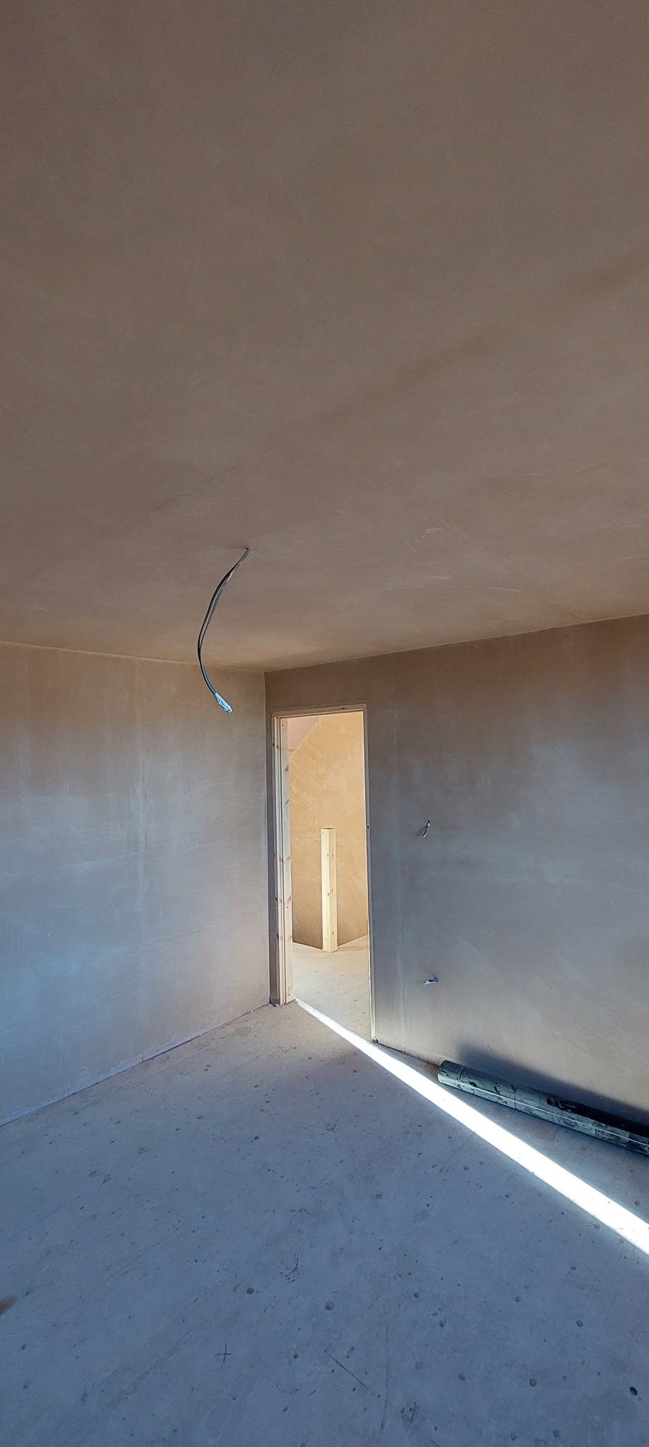 Wnętrze pokoju w stanie surowym po nałożeniu gładzi, widoczna wystająca instalacja elektryczna, otwór drzwiowy z widoczną konstrukcją w kolejnym pomieszczeniu, oraz narzędzie na podłodze.