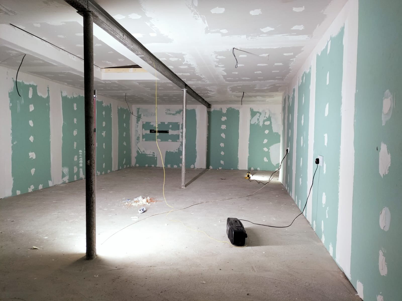 Surowe wnętrze w trakcie remontu: ściany pokryte płytami karton gips z widocznymi łączeniami do szpachlowania, metalowe podpory stropu, rozproszone kable elektryczne i narzędzia, betonowa podłoga...