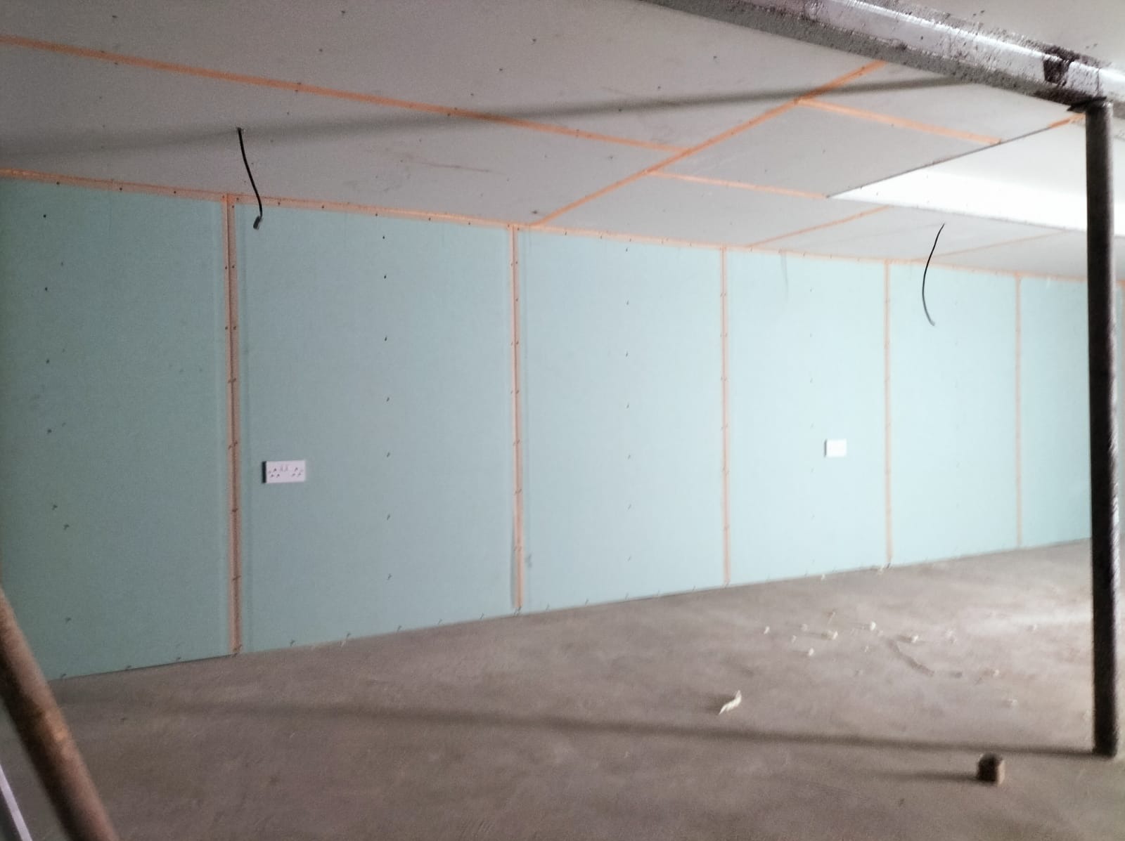 Wnętrze w trakcie prac wykończeniowych: surowe ściany z płyt kartonowo-gipsowych, widoczne łączenia i gniazdka elektryczne, betonowa podłoga z resztkami materiałów budowlanych, instalacja pod sufitem.