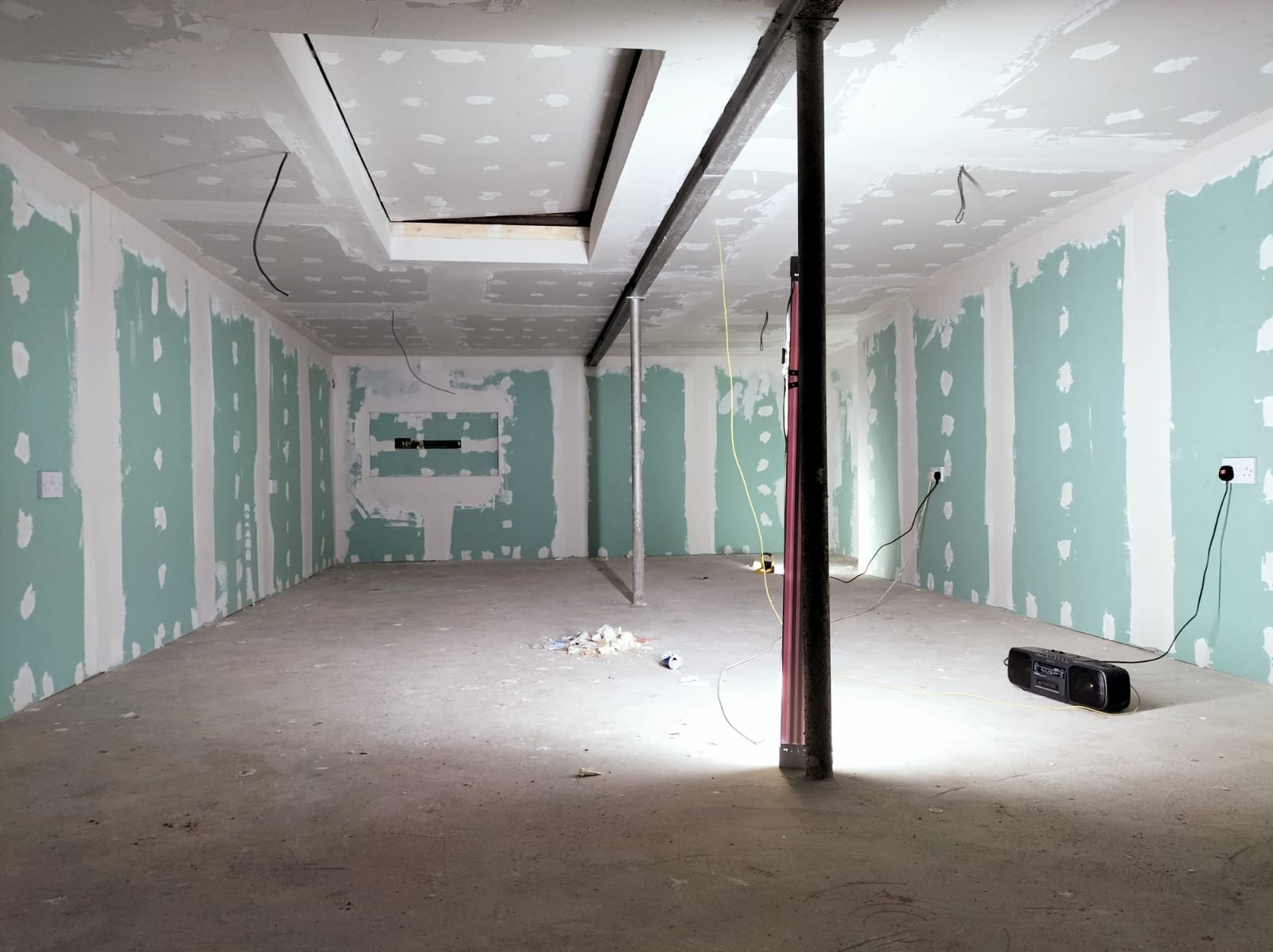 Surowe wnętrze w trakcie prac wykończeniowych: ściany z płyt gipsowo-kartonowych częściowo pokryte szpachlą, widoczne łączenia i śruby, metalowe podpory stropu, przewody elektryczne zwisające...