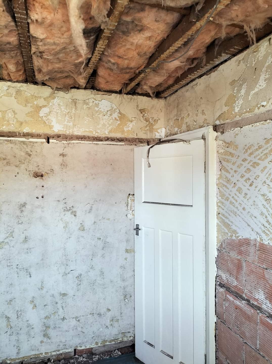 Pokój w trakcie remontu: odsłonięta izolacja stropu, fragmenty starej farby na ścianach, białe drzwi z kablem zwisającym nad górną krawędzią.