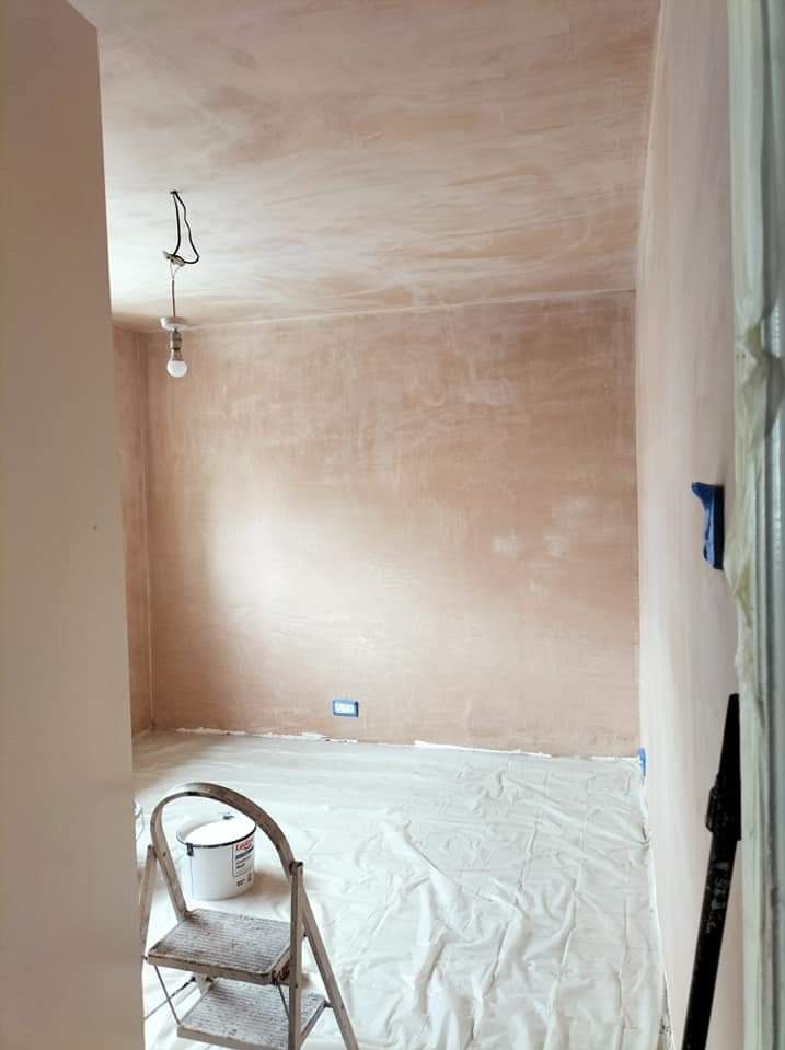Pokój przygotowany do malowania: ściany po wstępnym wyrównaniu, podłoga zabezpieczona folią, drabina z wiadrem w rogu, wisząca żarówka.