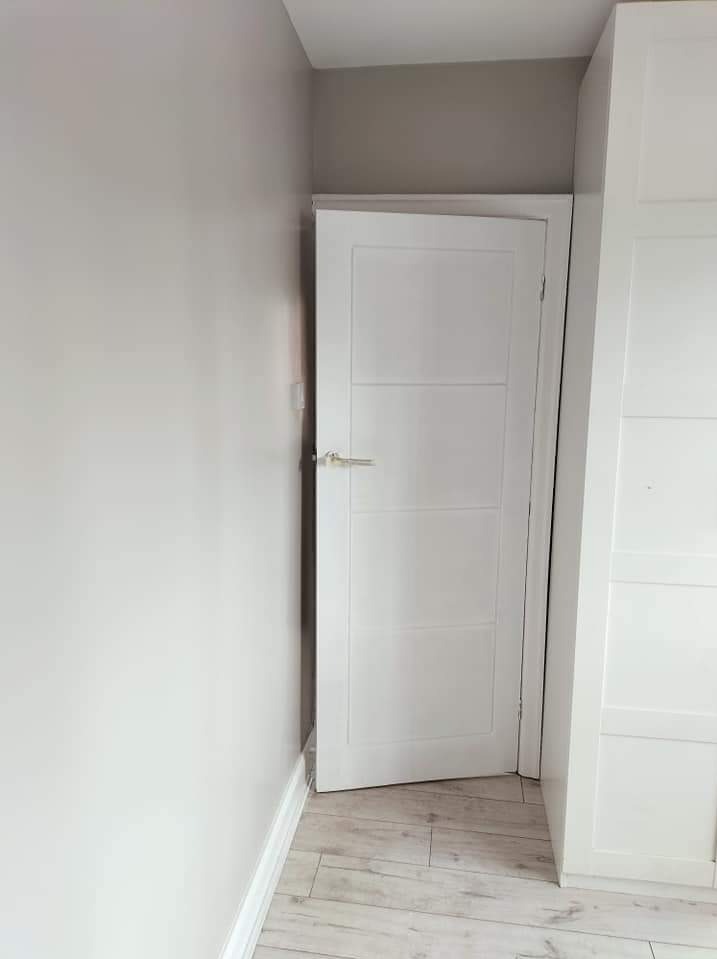 Wnętrze po remoncie: białe, częściowo otwarte drzwi z poziomymi panelami, srebrna klamka, szara ściana pomalowana farbą w odcieniach szarości, podłoga z paneli imitujących jasne drewno, biała szafa...