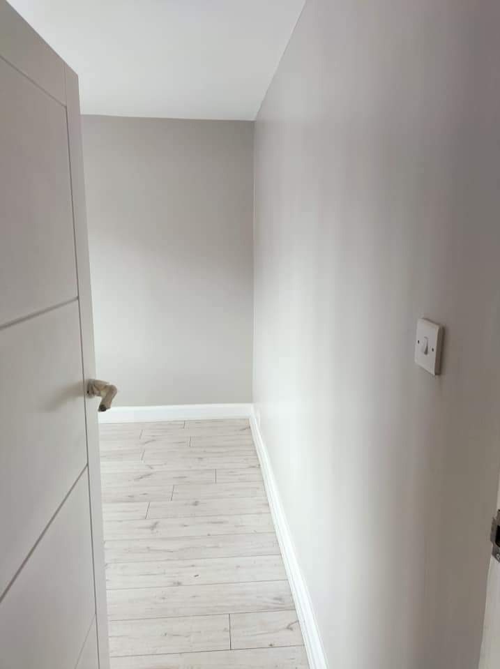 Korytarz po odświeżeniu, ściany pomalowane na jasny odcień szarości, białe listwy przypodłogowe, panele podłogowe imitujące bielone drewno, widoczny włącznik światła.