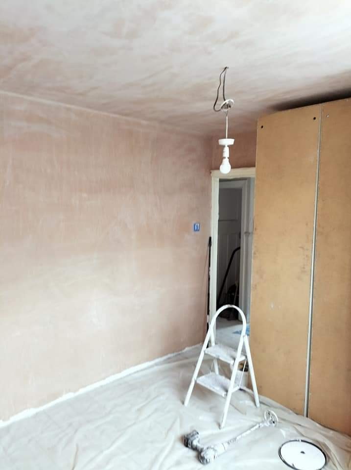 Pokój w trakcie szpachlowania: ściany pokryte świeżą warstwą szpachli, podłoga zabezpieczona folią malarską, widoczna biała drabina malarska i wisząca żarówka bez klosza.