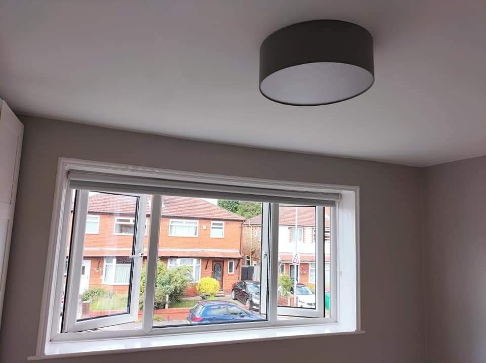 Świeżo pomalowane wnętrze pokoju z białym oknem PCV, częściowo otwartym, widok na ulicę z ceglanymi domami i samochodami, szary abażur lampy sufitowej.