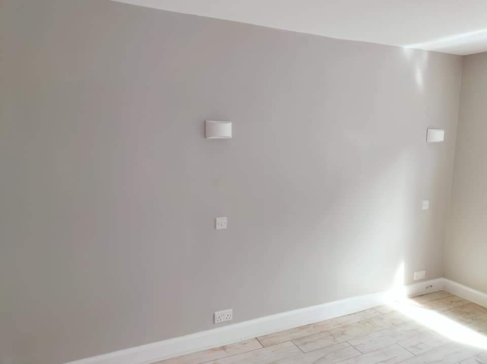 Świeżo pomalowane wnętrze pokoju w odcieniach szarości, widoczne gniazdka elektryczne i lampa ścienna, wykończone listwy przypodłogowe i panele podłogowe imitujące drewno.