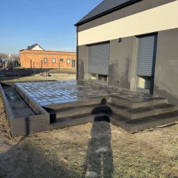 FBK BRUK Bartosz Haremza - Nowoczesny taras z ciemnej kostki brukowej z niskim murkiem oporowym, widoczny cień osoby fotografującej, w tle budynek z szarymi roletami.