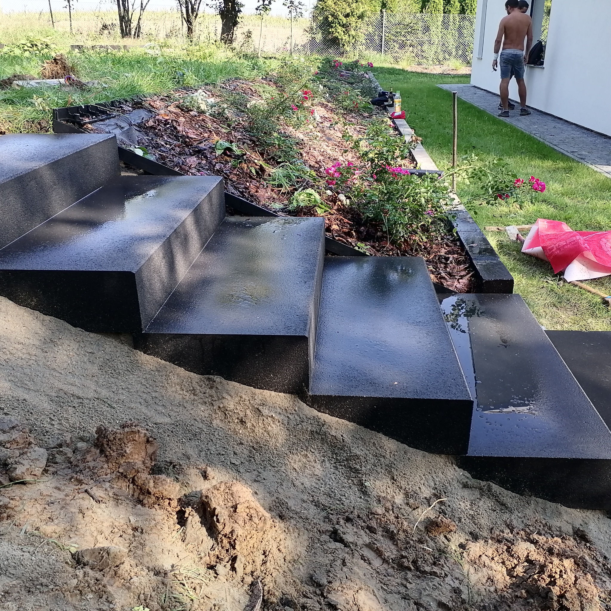 Schody zewnętrzne z czarnej kostki brukowej prowadzące do ogrodu z rabatą kwiatową, w tle budynek mieszkalny i osoby.