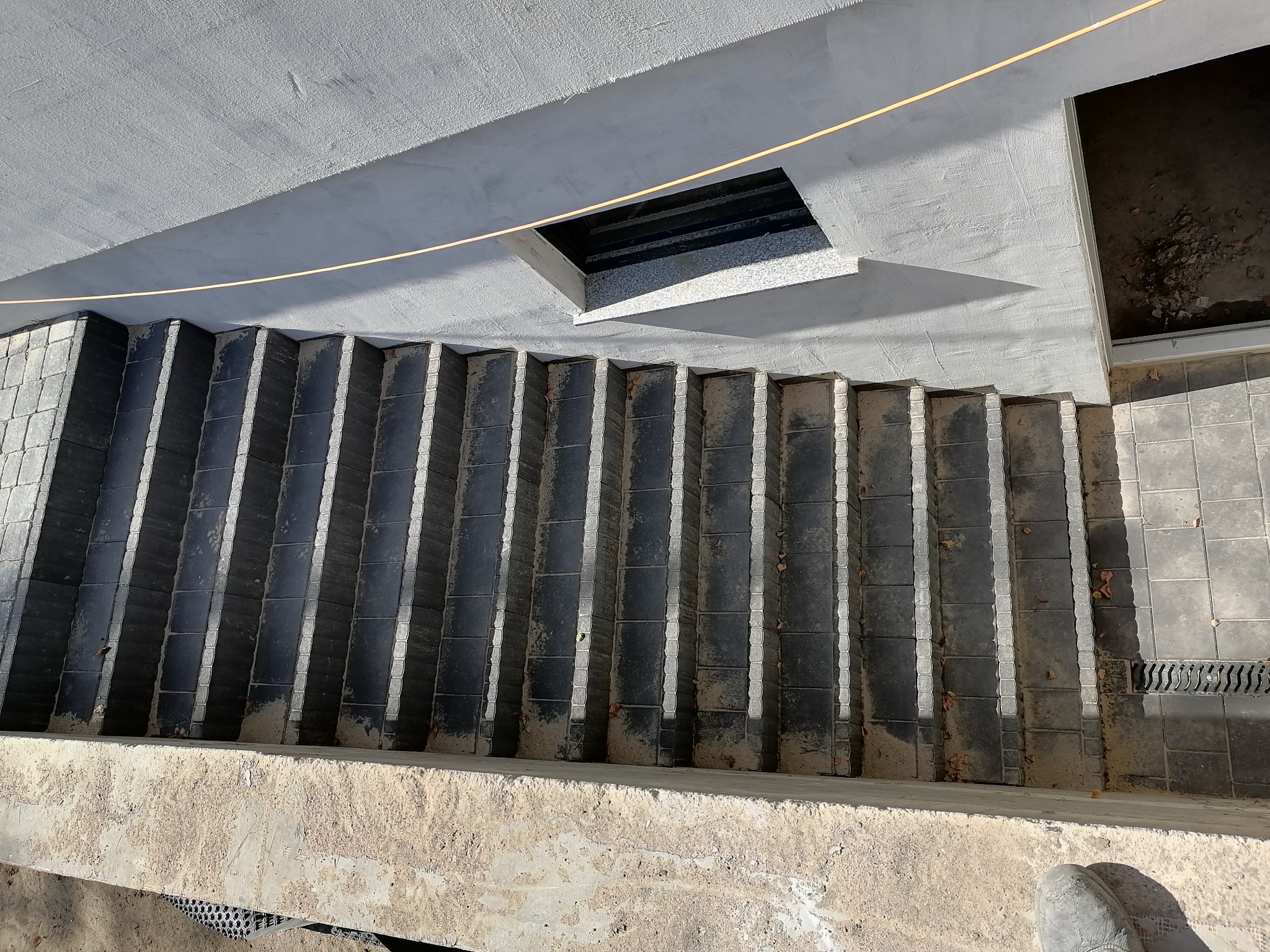 Schody wyłożone ciemną kostką brukową z jasnymi fugami, widok z góry, betonowy sufit z otworem wentylacyjnym w tle.