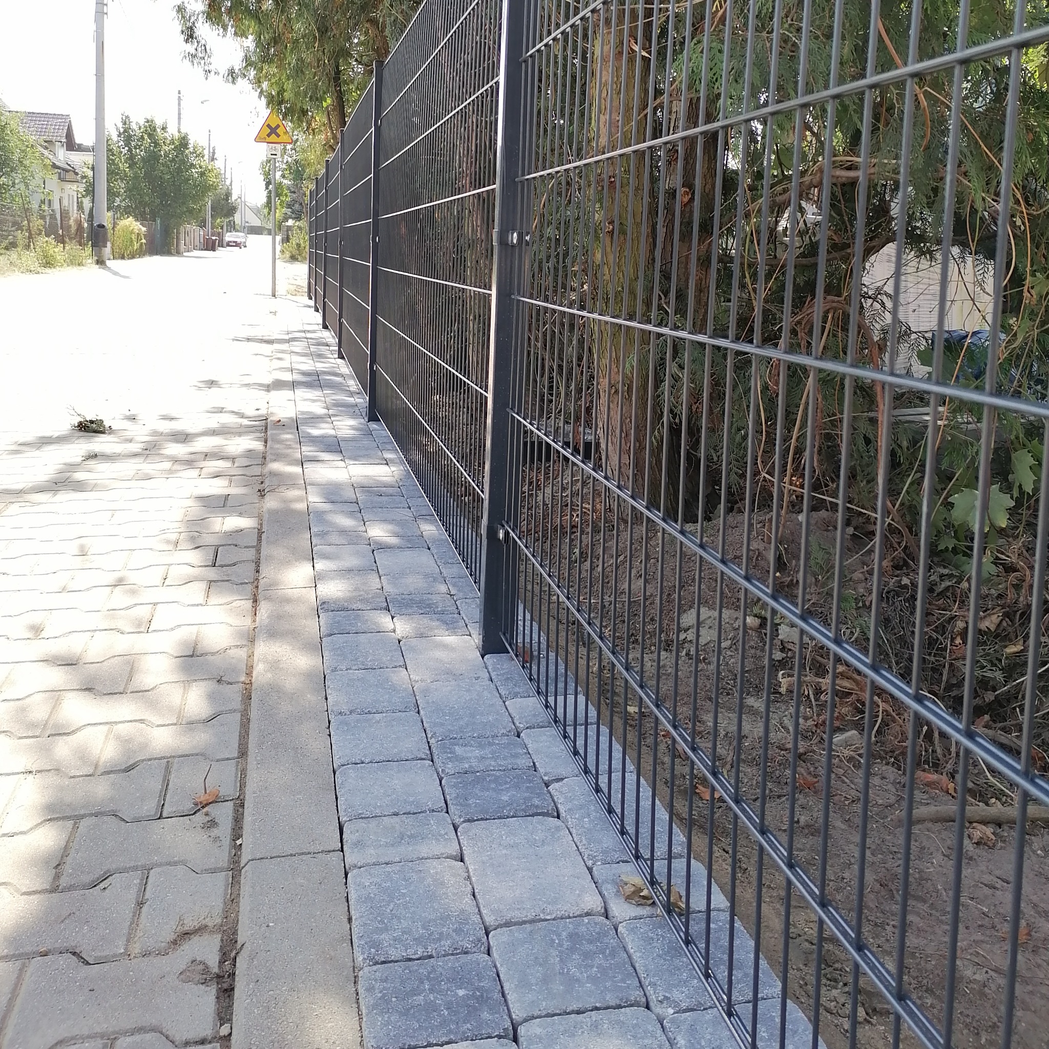Długi, prosty odcinek nowoczesnego, metalowego ogrodzenia panelowego w kolorze grafitowym, zamontowanego wzdłuż chodnika z szarej kostki brukowej. W tle widoczna ulica i drzewa.