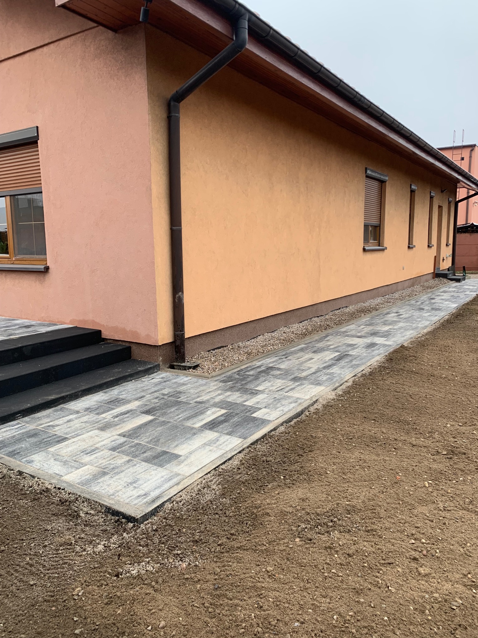 Świeżo ułożony chodnik z szaro-białej kostki brukowej prowadzący wzdłuż ściany domu o brzoskwiniowym odcieniu, z widocznymi schodami wejściowymi i przygotowanym podłożem z ziemi.