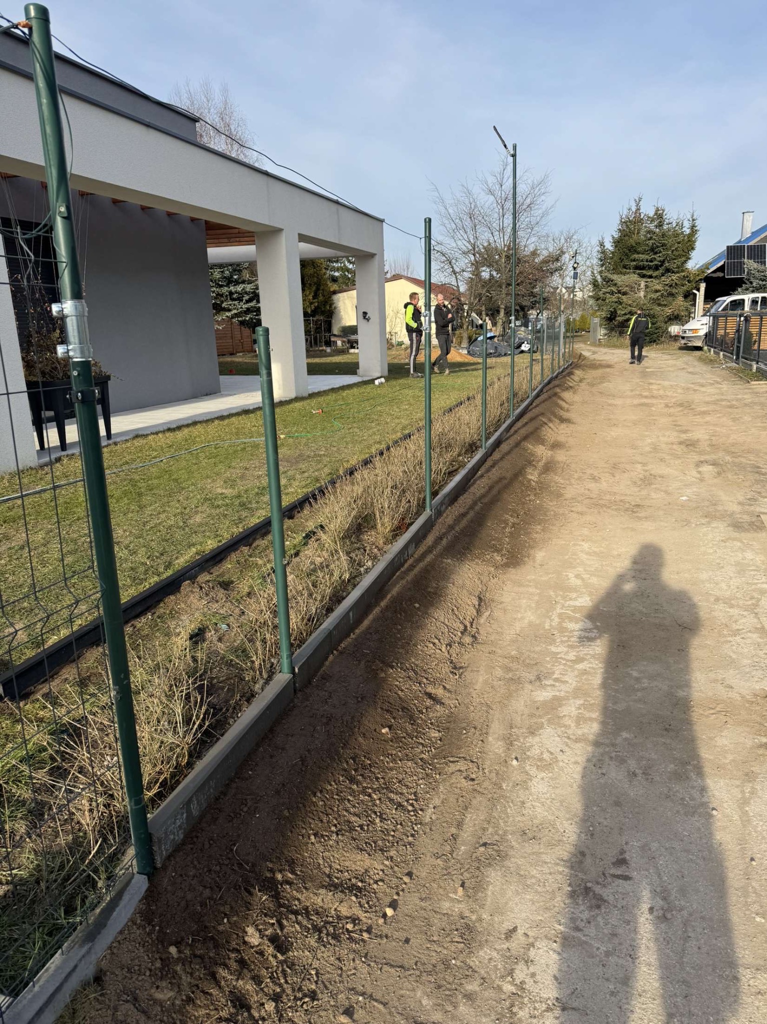 Montaż ogrodzenia panelowego z zielonymi słupkami na betonowej podmurówce, widoczni pracownicy w odblaskowych kamizelkach, w tle dom z ogrodem.