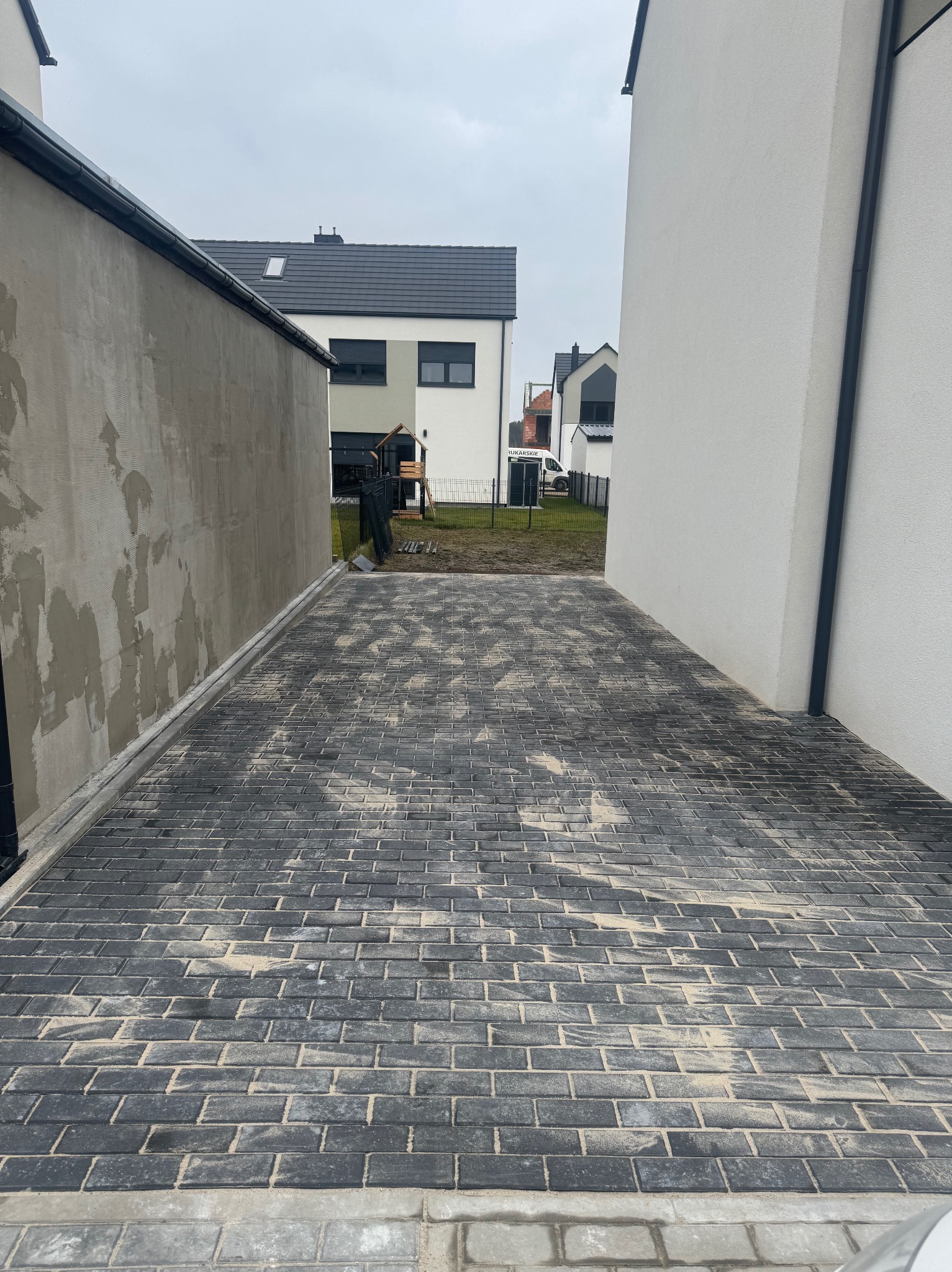 Ułożona, szara kostka brukowa na podjeździe między dwoma budynkami, z widocznym pracownikiem w oddali wyrównującym piasek.