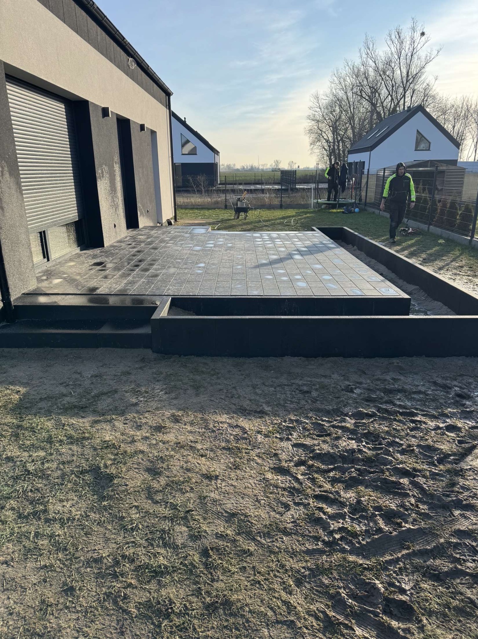 Nowoczesny taras z ciemnej kostki brukowej, zlokalizowany przy domu jednorodzinnym we Wrześni. Widoczne schody prowadzące na taras oraz przygotowane obrzeże. W tle pracownicy i krajobraz podmiejski.