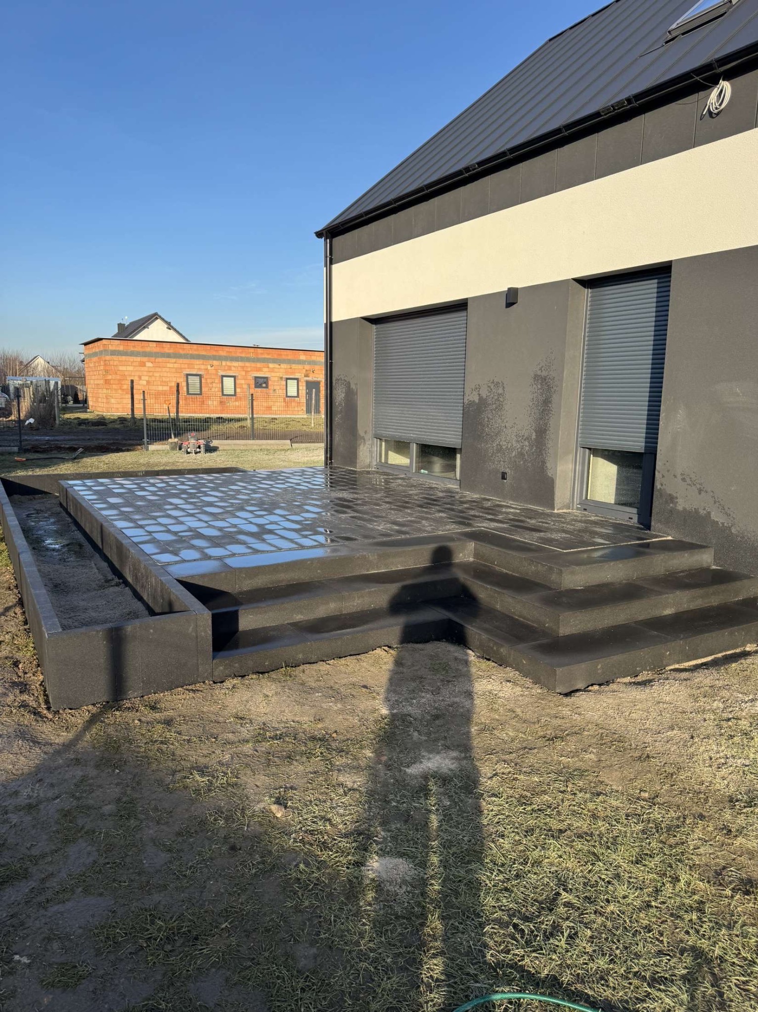 Nowoczesny taras z ciemnej kostki brukowej z niskim murkiem oporowym, widoczny cień osoby fotografującej, w tle budynek z szarymi roletami.