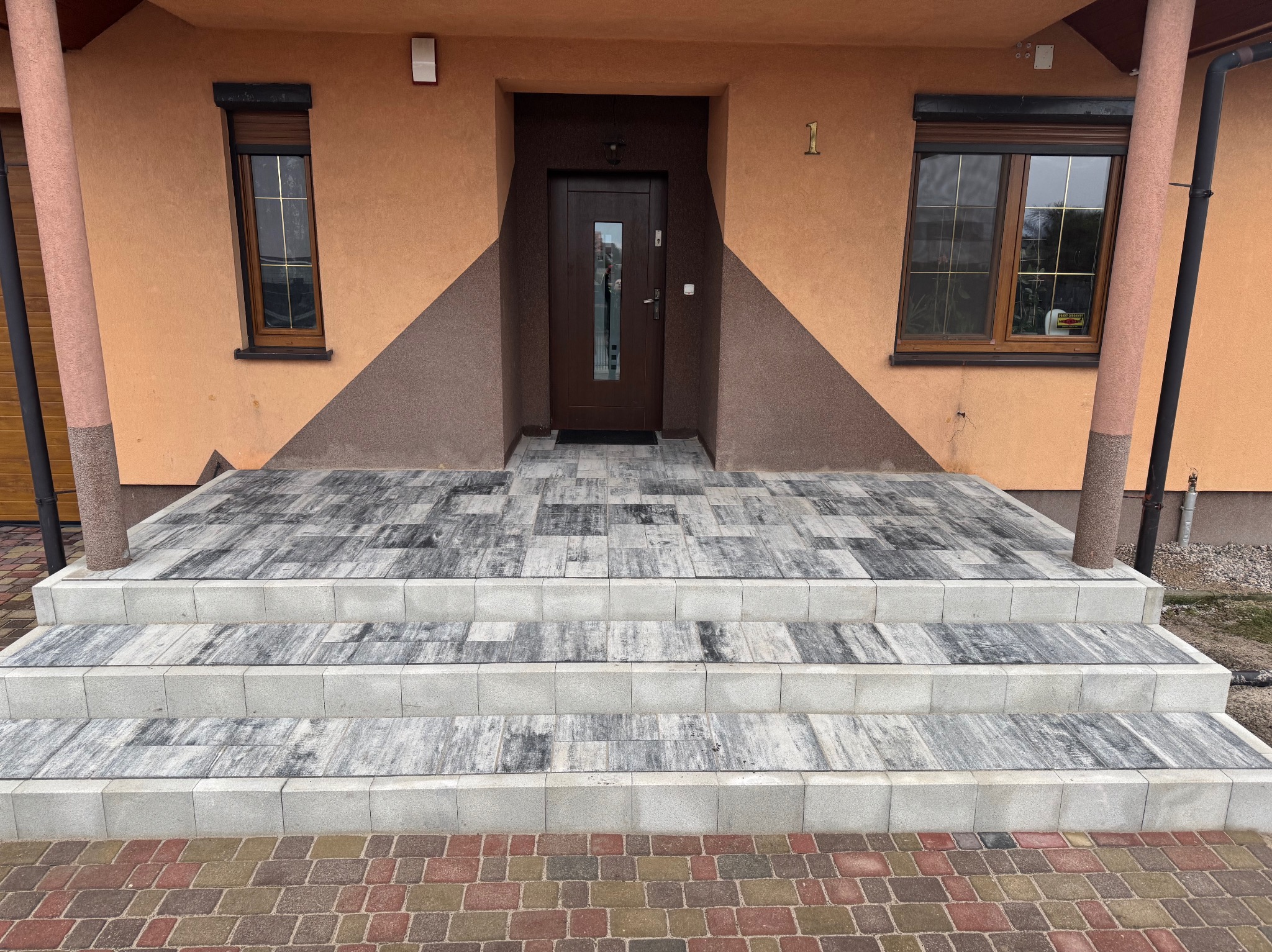 Schody wejściowe wyłożone szarą kostką brukową na zewnątrz budynku z brązowymi oknami i drzwiami.