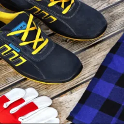 Niebieskie buty robocze z żółtymi sznurówkami marki Roma, czerwono-białe rękawice robocze Pruspol Brook i flanelowa koszula w niebiesko-czarną kratę leżą na drewnianym podłożu.