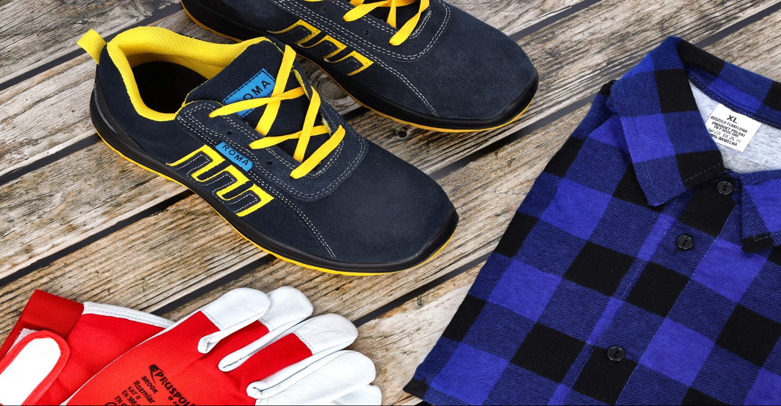 Niebieskie buty robocze z żółtymi sznurówkami marki Roma, czerwono-białe rękawice robocze Pruspol Brook i flanelowa koszula w niebiesko-czarną kratę leżą na drewnianym podłożu.