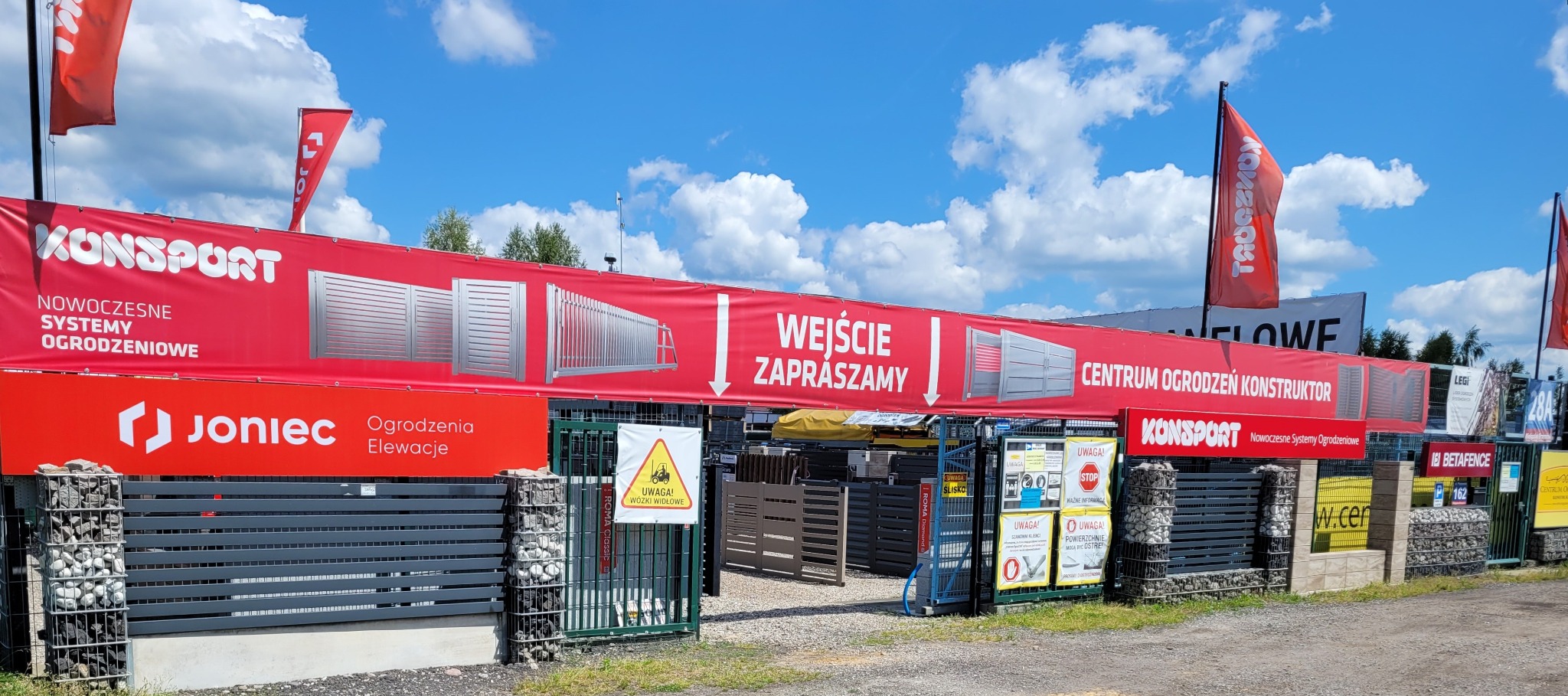 Ekspozycja systemów ogrodzeniowych i bram wjazdowych w centrum ogrodzeń, z widocznymi panelami, ogrodzeniami metalowymi i reklamami firm Joniec i Konsport, pod błękitnym niebem z chmurami.
