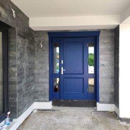 REVERS Studio Drzwi - Łukasz Niemiec - Niebieskie drzwi wejściowe z bocznymi przeszkleniami, zamontowane w budynku z elewacją z szarego kamienia, widoczne ślady prac wykończeniowych na posadzce.