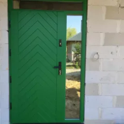 Świeżo zamontowane, zielone drzwi wejściowe z szybą, wzór w jodełkę, osadzone w surowym murze z betonu komórkowego. Widoczne przewody elektryczne i skrzynka narzędziowa.