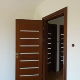 Częściowo otwarte, ciemnobrązowe drzwi wewnętrzne z poziomymi, białymi wstawkami na tle białej ściany i drewnianej podłogi; w tle widoczne kolejne drzwi w podobnym stylu.