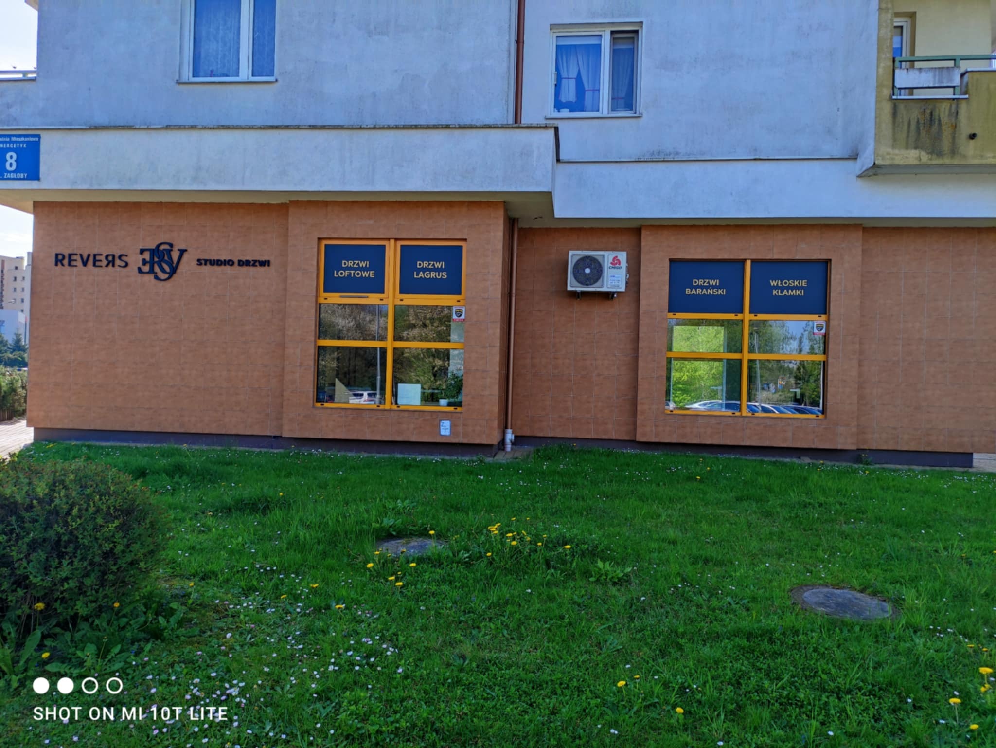 Fasada budynku z szyldem 'REVERS Studio Drzwi' i oknami wystawowymi prezentującymi różne modele drzwi, w tym 'Drzwi Loftowe', 'Drzwi Lagrus' oraz 'Drzwi Baranski' i 'Włoskie Klamki', z widoczną...