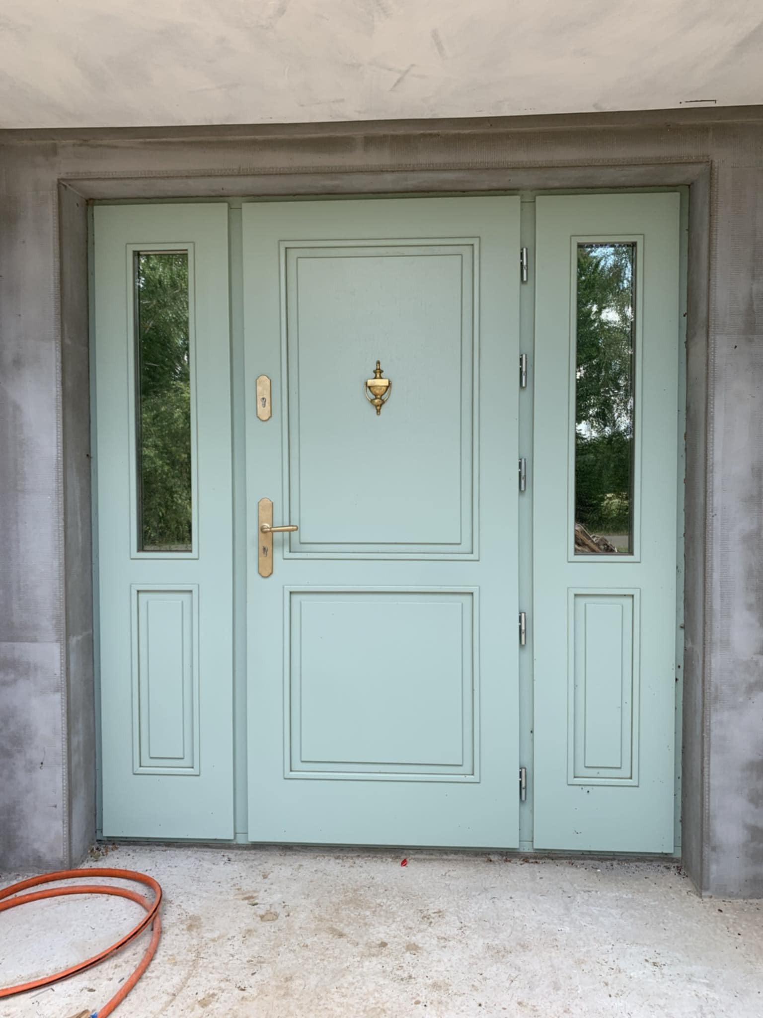 Jasnozielone drzwi wejściowe z bocznymi panelami szklanymi i złotym kołatką, osadzone w surowej, betonowej ramie, widoczne pomarańczowe kable na podłodze.