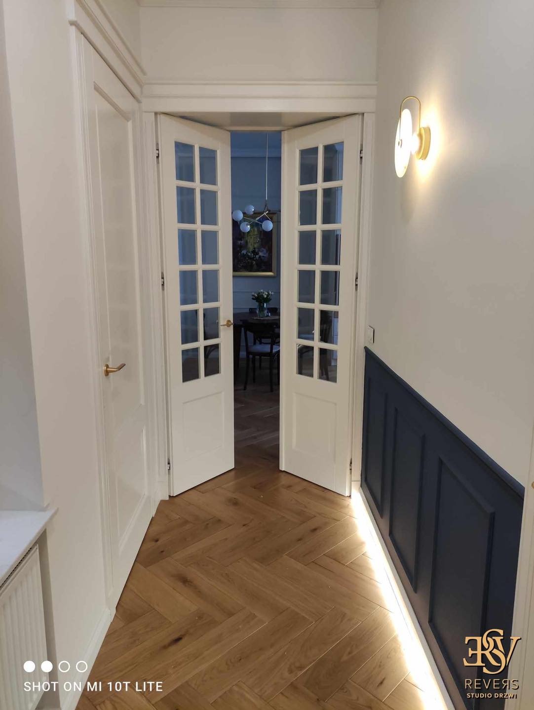 Korytarz z parkietem w jodełkę i otwartymi dwuskrzydłowymi drzwiami z przeszkleniami, widok na wnętrze pokoju z żyrandolem i obrazem, oświetlenie ścienne i listwa przypodłogowa z podświetleniem LED.