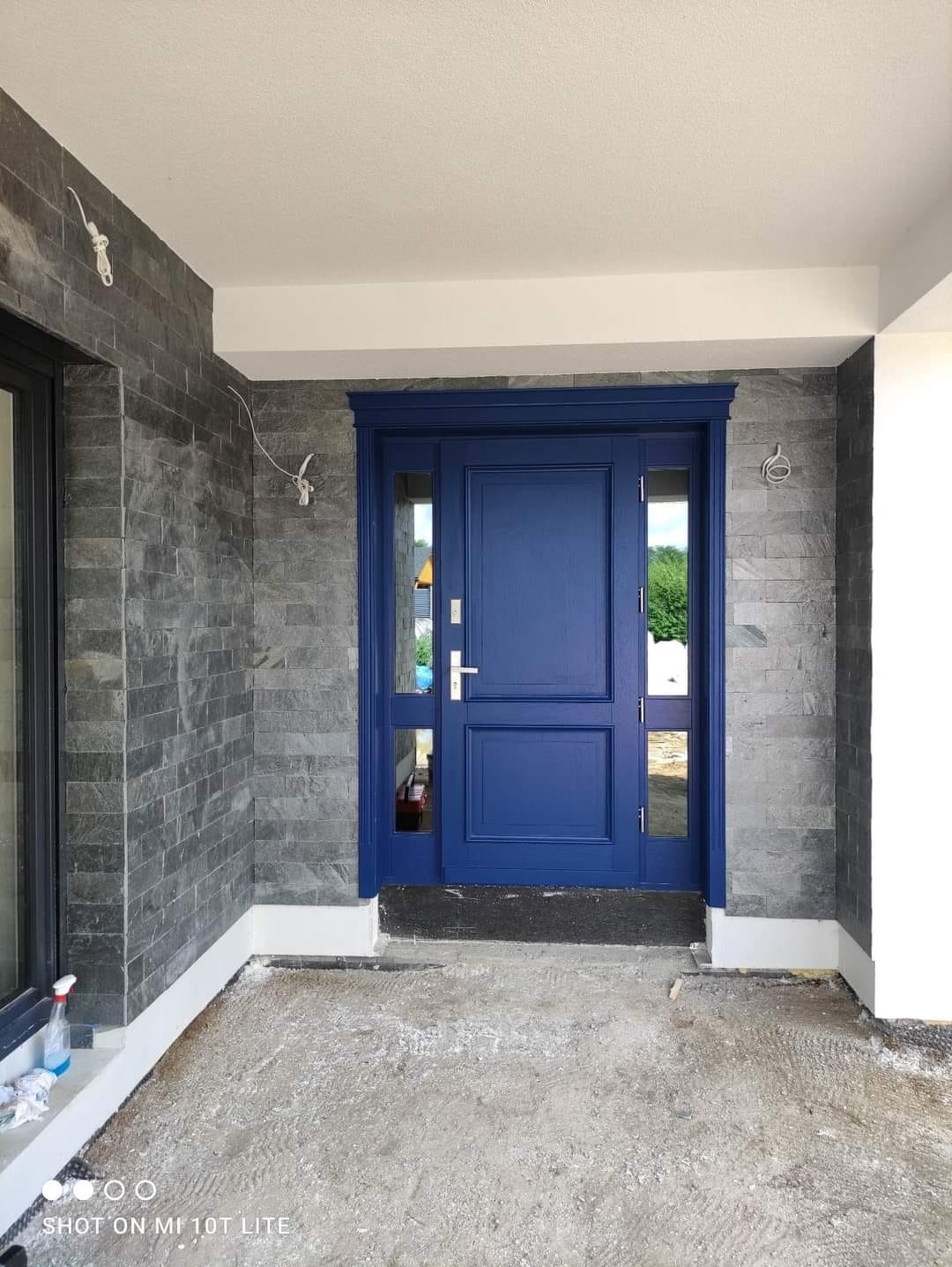 Niebieskie drzwi wejściowe z bocznymi przeszkleniami, zamontowane w budynku z elewacją z szarego kamienia, widoczne ślady prac wykończeniowych na posadzce.