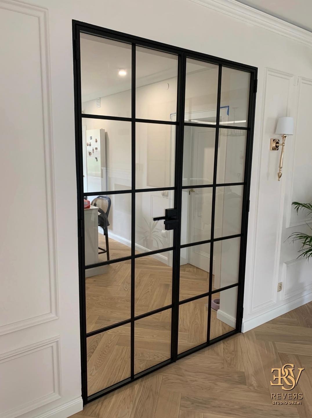 Dwuskrzydłowe drzwi loftowe z czarnymi szprosami i przezroczystym szkłem, widok na wnętrze z parkietem w jodełkę i białą ścianą z dekoracyjną listwą, oświetlone złotym kinkietem.