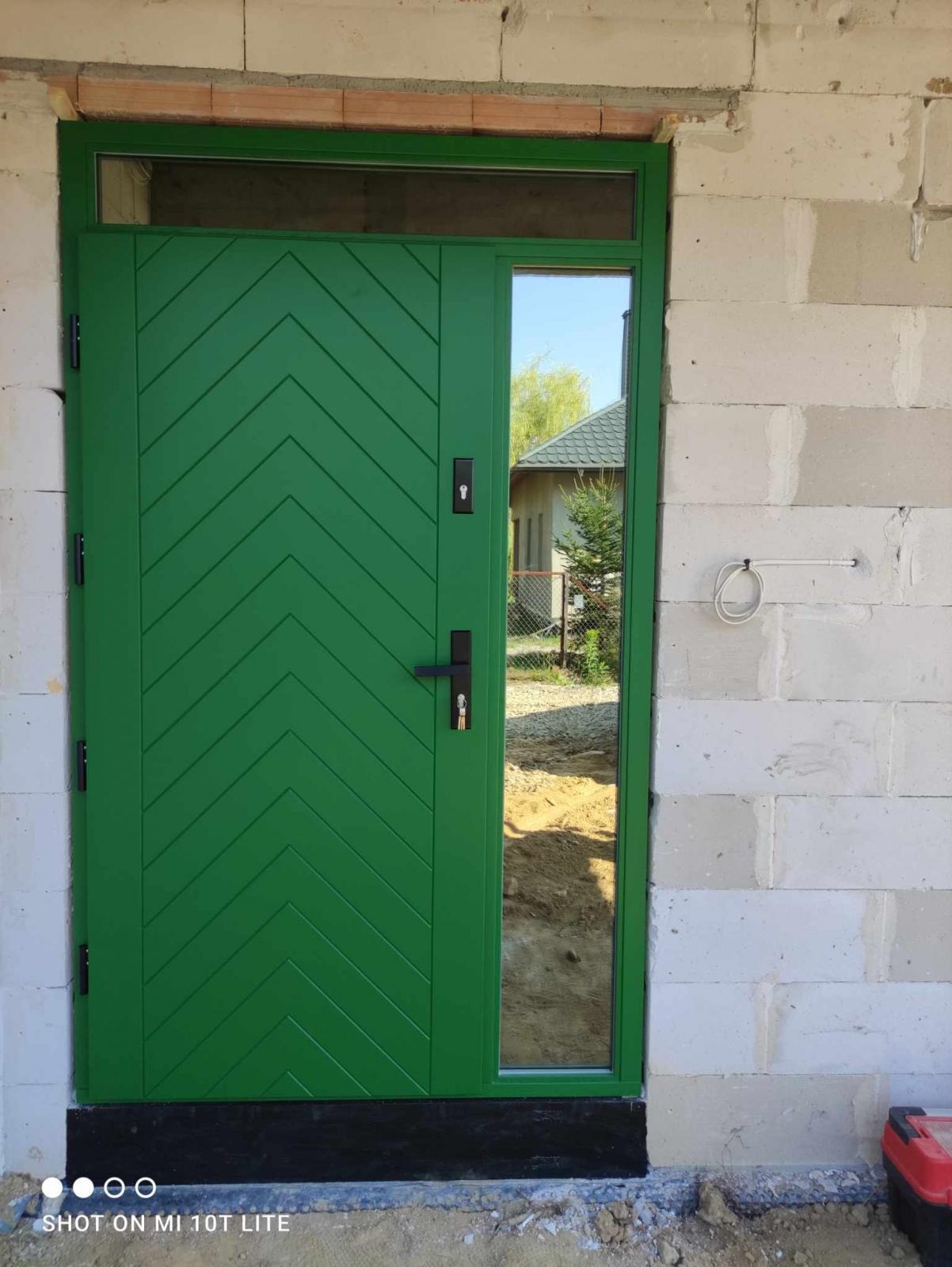 Świeżo zamontowane, zielone drzwi wejściowe z szybą, wzór w jodełkę, osadzone w surowym murze z betonu komórkowego. Widoczne przewody elektryczne i skrzynka narzędziowa.