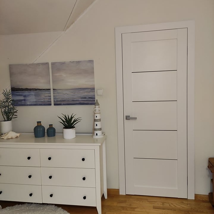 Białe drzwi wewnętrzne z czterema poziomymi wgłębieniami, zamontowane w pokoju z komodą, obrazami o tematyce morskiej i drewnianą podłogą.