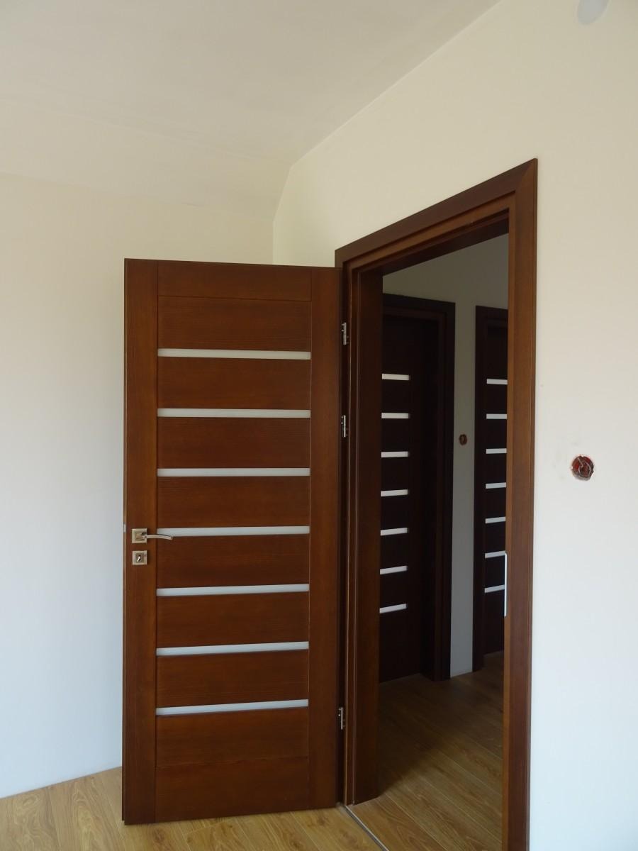 Częściowo otwarte, ciemnobrązowe drzwi wewnętrzne z poziomymi, białymi wstawkami na tle białej ściany i drewnianej podłogi; w tle widoczne kolejne drzwi w podobnym stylu.