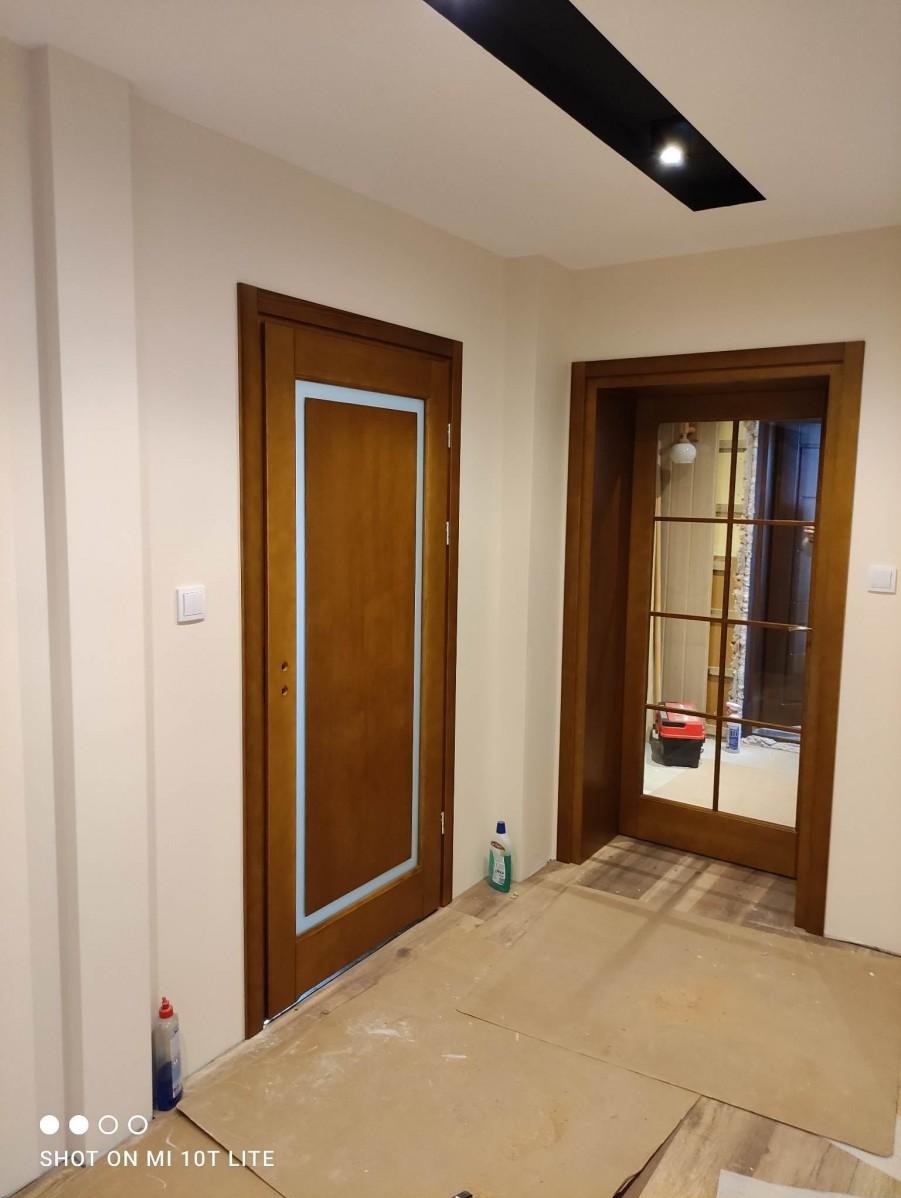 Dwa rodzaje drzwi wewnętrznych: pełne drewniane z niebieską wstawką oraz przeszklone z drewnianą ramą, widoczne w korytarzu podczas remontu, zabezpieczona podłoga, butelki z płynami na podłodze.