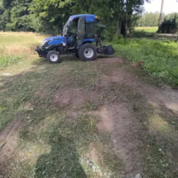 Niebieski traktor z otwartymi drzwiami kabiny, stojący na częściowo skoszonej trawie na skraju pola, widoczne ślady opon, tło stanowią drzewa i budynek w oddali.