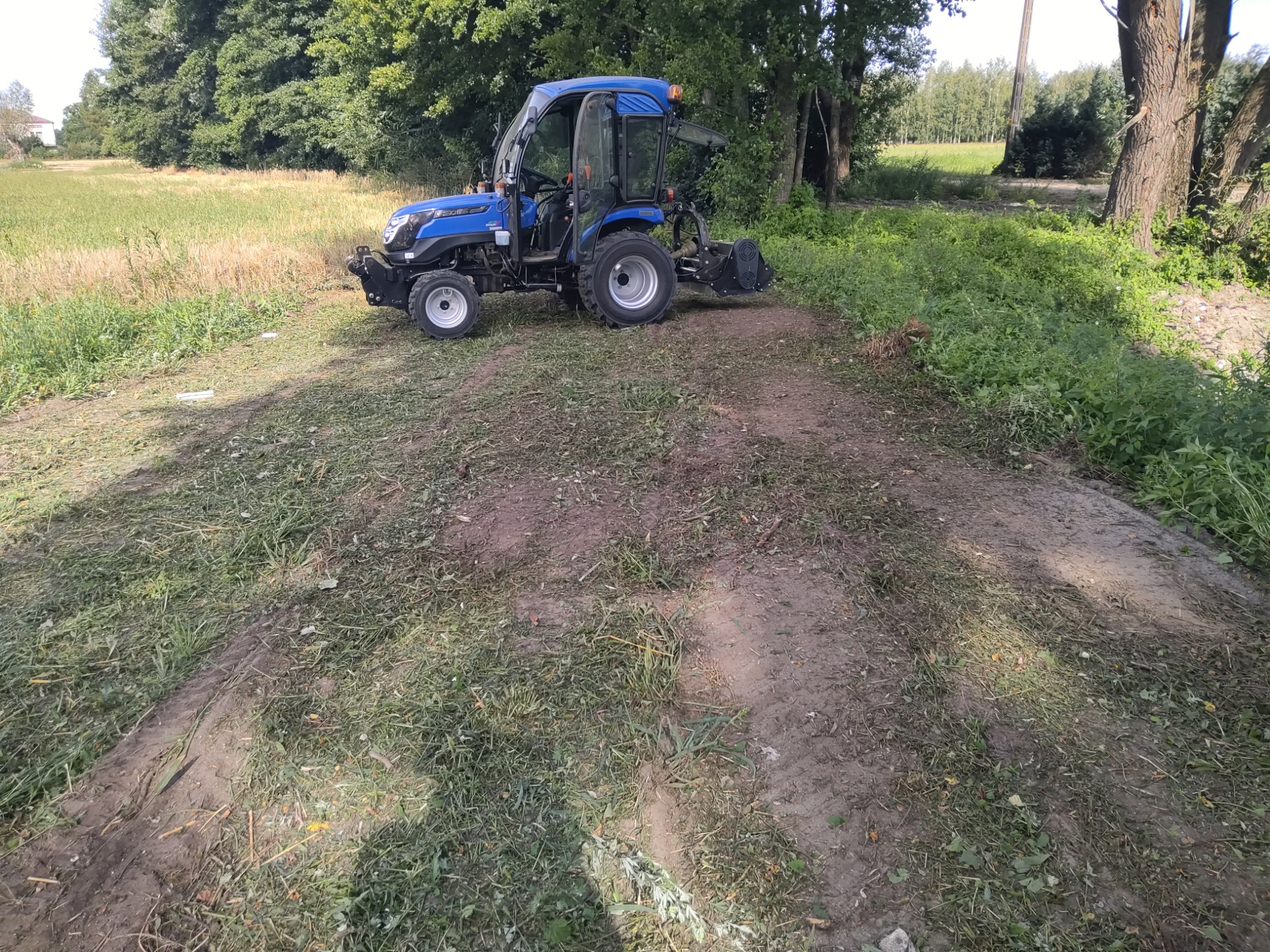 Niebieski traktor z otwartymi drzwiami kabiny, stojący na częściowo skoszonej trawie na skraju pola, widoczne ślady opon, tło stanowią drzewa i budynek w oddali.