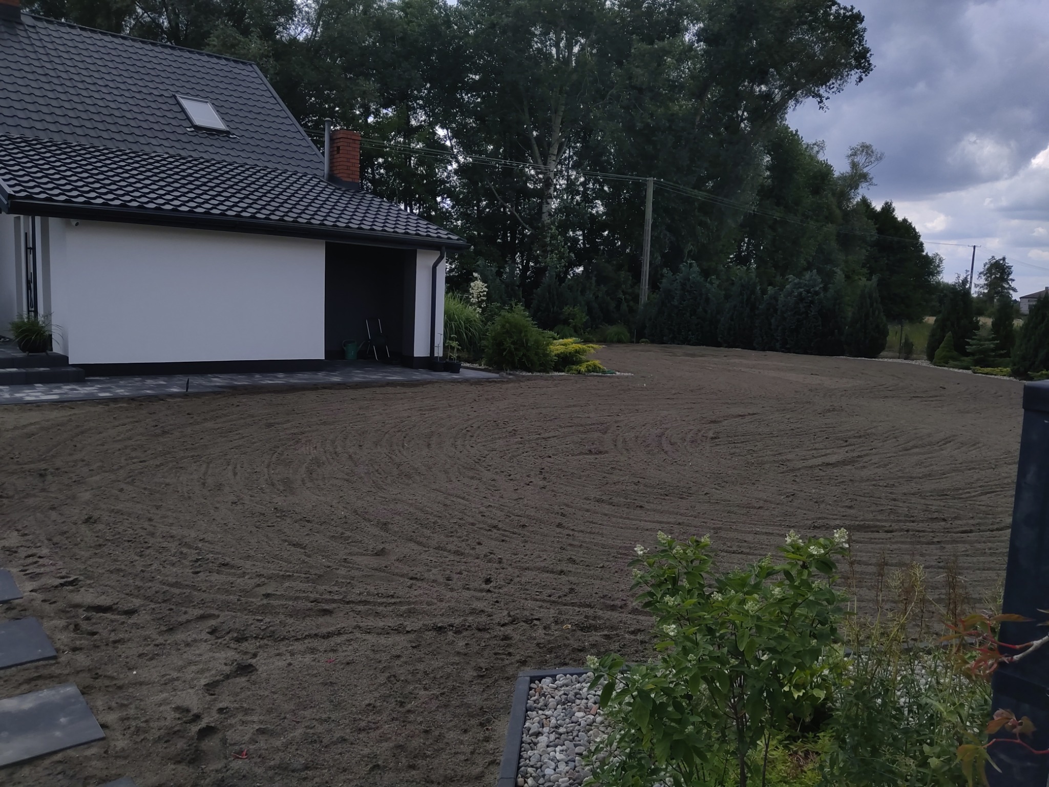 Przygotowany teren pod trawnik po niwelacji, widoczne ślady po maszynach, nowoczesny dom z ciemną dachówką w tle, bujna roślinność w ogrodzie.