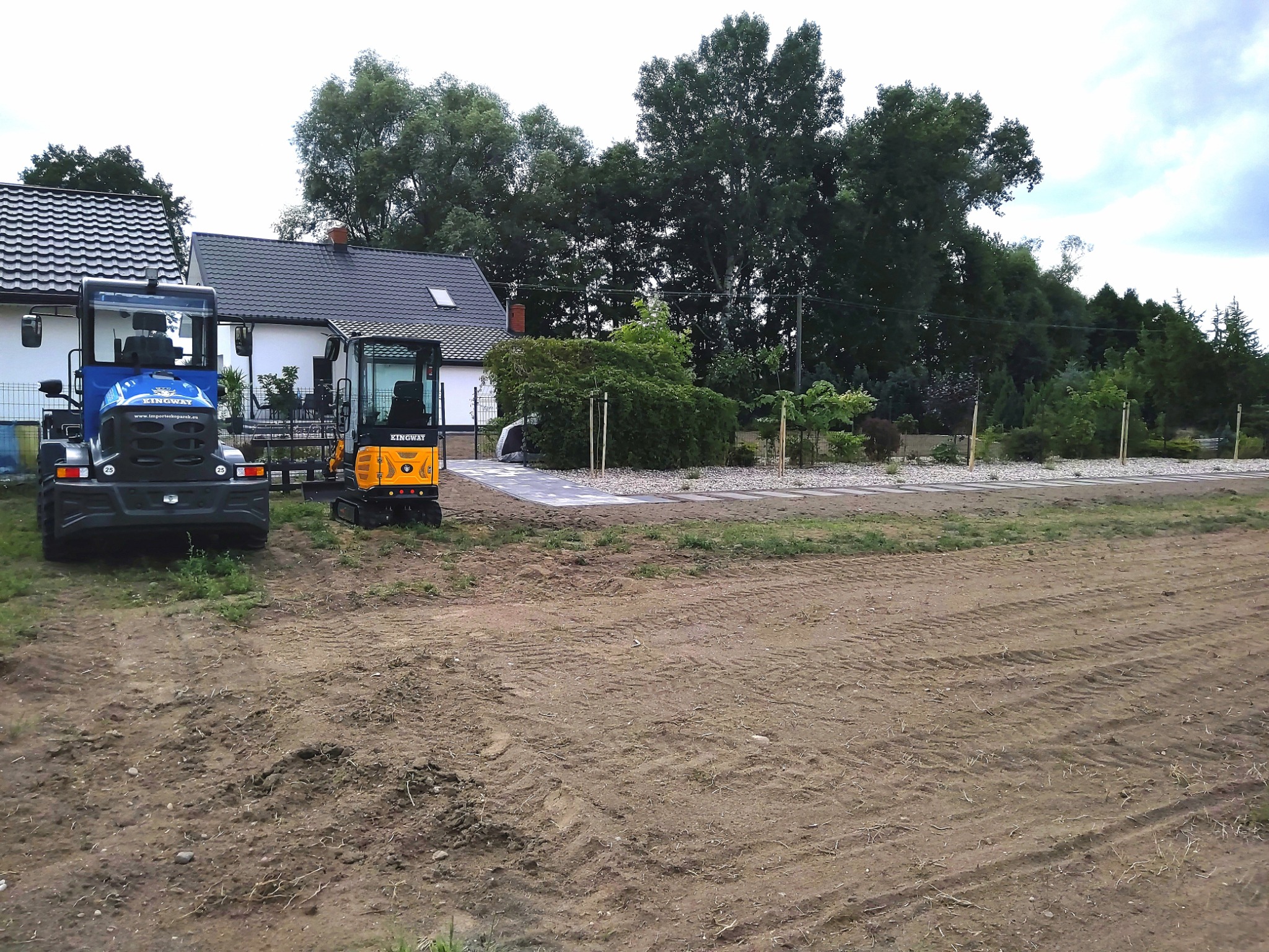 Nierówny teren po robotach ziemnych z dwoma małymi koparkami Kingway, niebieską i żółtą, na tle białego domu z ciemnym dachem i zieleni drzew