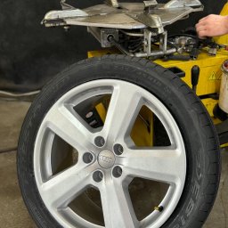 CASTROCAR SPÓŁKA Z OGRANICZONĄ ODPOWIEDZIALNOŚCIĄ - Srebrna felga Audi z oponą Debica na tle montażownicy do opon w warsztacie w Warszawie. Widoczny fragment dłoni pracownika.