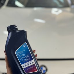 CASTROCAR SPÓŁKA Z OGRANICZONĄ ODPOWIEDZIALNOŚCIĄ - Oryginalny olej silnikowy BMW Twin Power Turbo SAE 5W-30 Longlife-04, trzymany w dłoni na tle białego samochodu BMW, widoczne logo BMW na opakowaniu i na samochodzie.