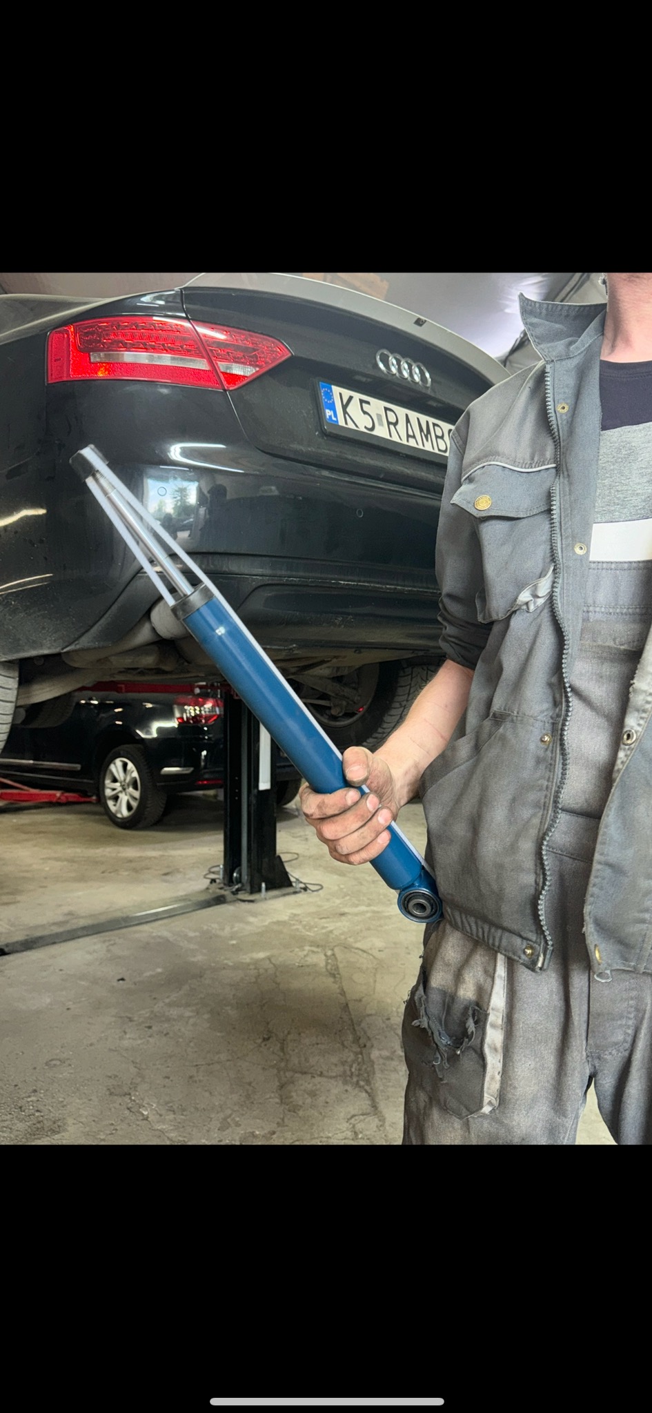 Mechanik w szarym kombinezonie trzyma nowy, niebieski amortyzator na tle podnośnika z podniesionym czarnym Audi z widoczną tablicą rejestracyjną K5 RAMBO.