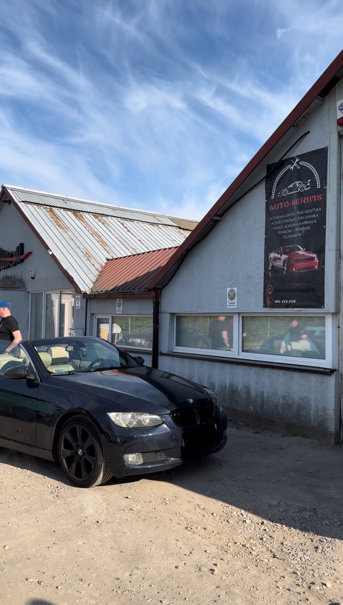 Dwa czarne BMW zaparkowane przed warsztatem samochodowym z banerem reklamowym usług: wymiana opon, diagnostyka komputerowa, mechanika samochodowa, naprawa silników, spawanie, tłumiki. Widoczne okna...